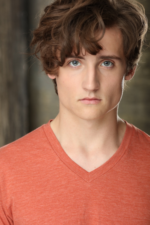 A young person with curly hair and blue eyes, wearing an orange V-neck shirt, poses against a blurred background.