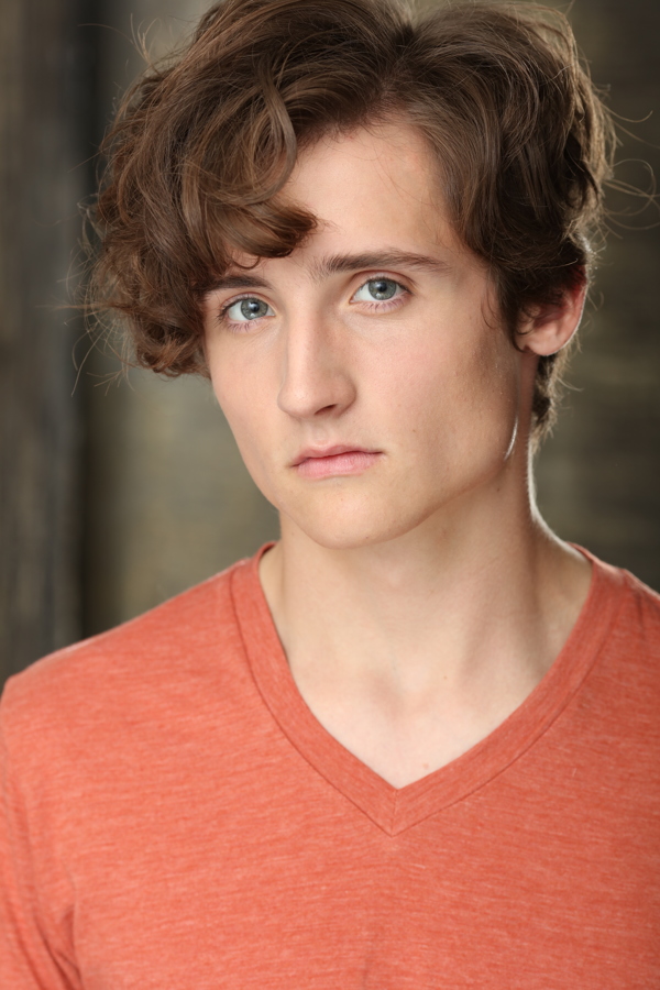 A young person with curly hair wearing an orange v-neck shirt, looking serious against a blurred background.