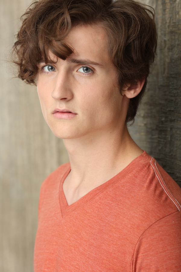 A young person with curly hair, wearing an orange v-neck shirt, poses against a textured background.