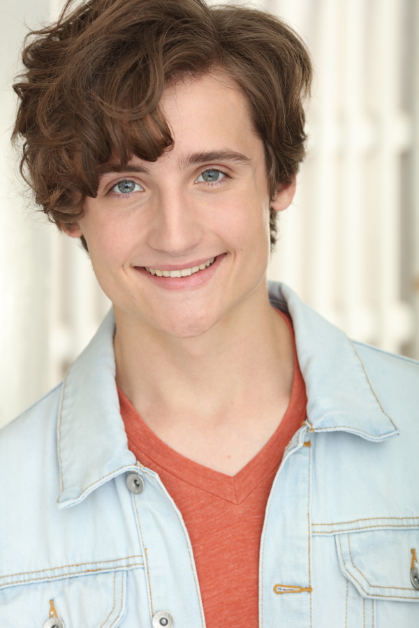 A young person with curly hair and blue eyes smiling, wearing a denim jacket over a red shirt.