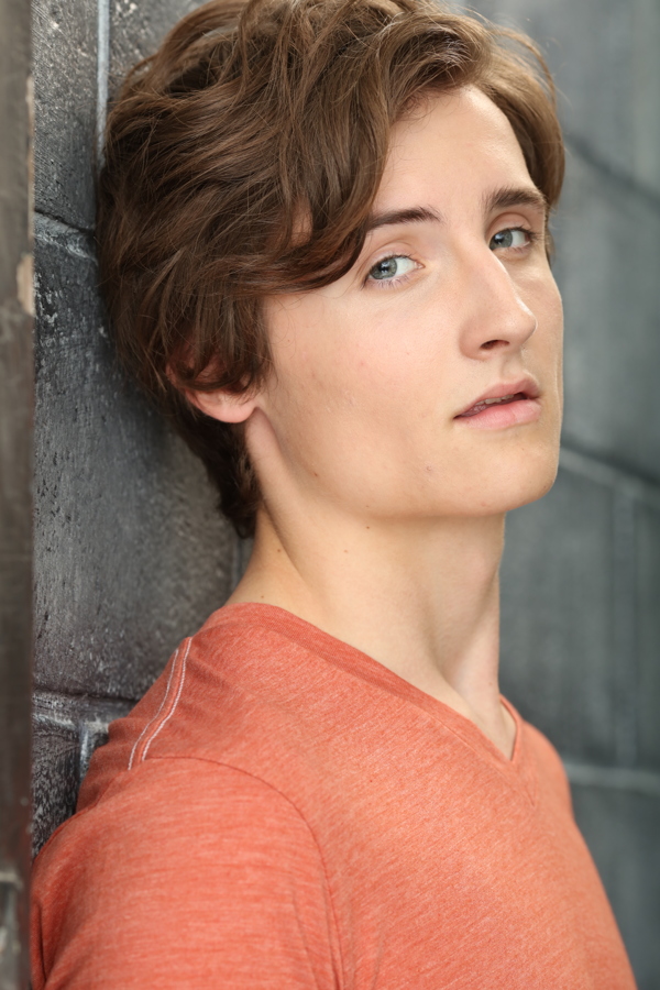 A young person with wavy hair poses against a gray wall, wearing an orange shirt, looking thoughtfully at the camera.