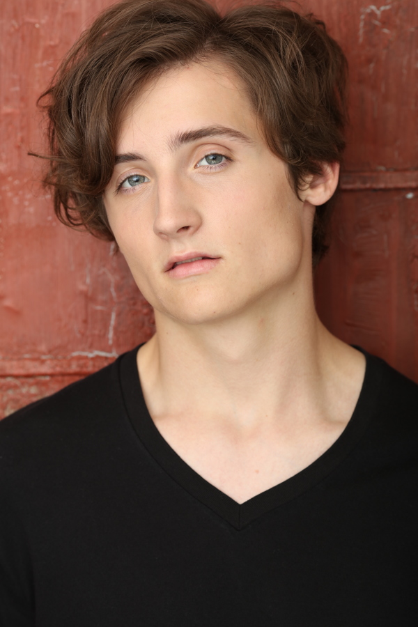 A young person with tousled hair and blue eyes, wearing a black V-neck shirt, poses against a red wall.