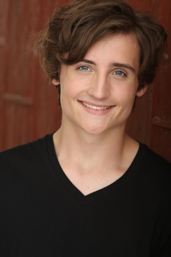 A smiling young person with short, wavy hair and blue eyes, wearing a black V-neck shirt against a wooden background.