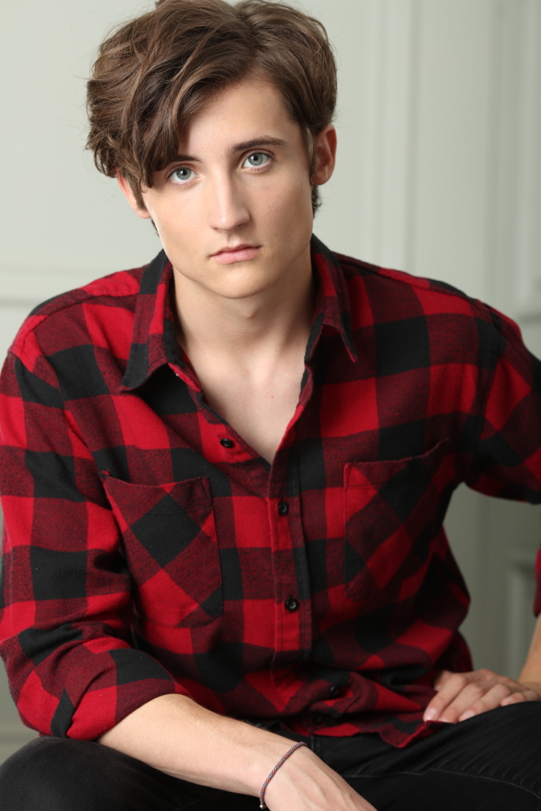 A young person with short wavy hair poses in a red and black checkered shirt, looking directly at the camera.