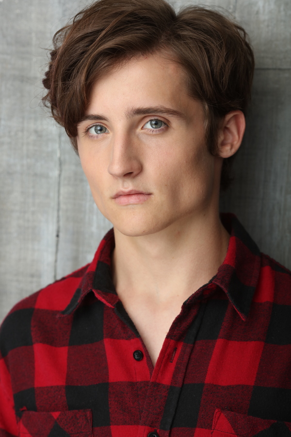 A young man with tousled hair wearing a red and black plaid shirt, looking directly at the camera against a gray background.