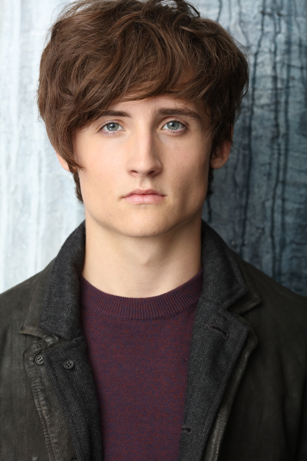 A young person with tousled hair and blue eyes, wearing a dark coat and a burgundy sweater.