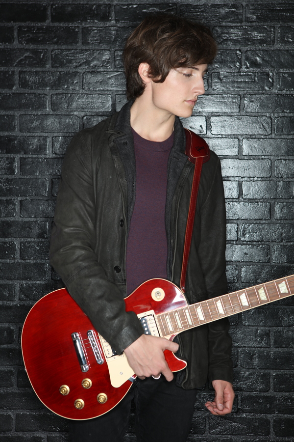 A young person stands against a black wall, holding a red electric guitar, dressed in casual attire.