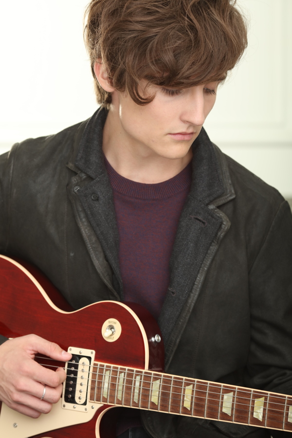 A young man with wavy hair plays a red electric guitar, wearing a dark jacket and looking thoughtfully down.