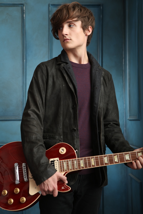 A young person with wavy hair is holding an electric guitar, wearing a black jacket against a blue backdrop.