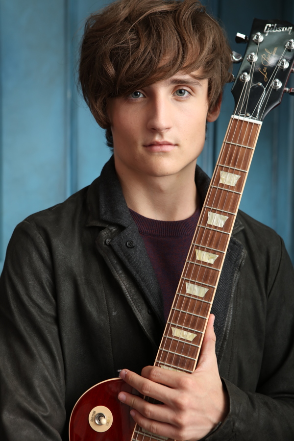 A young man with tousled hair holding a guitar, dressed in a black jacket against a blue background.